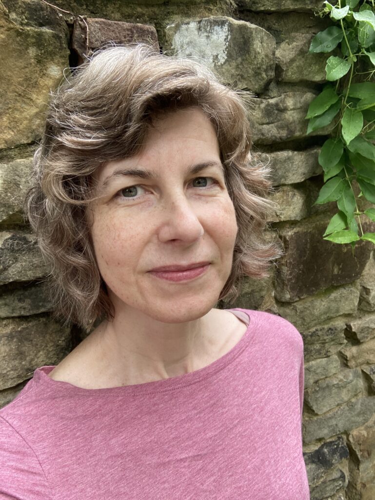 Woman with short brown hair and pink shirt pictured from chest up looking into the camera against a brick background.